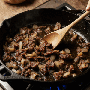 Carne seca desfiada sendo refogada com cebola em uma frigideira de ferro, mexida com uma colher de pau.