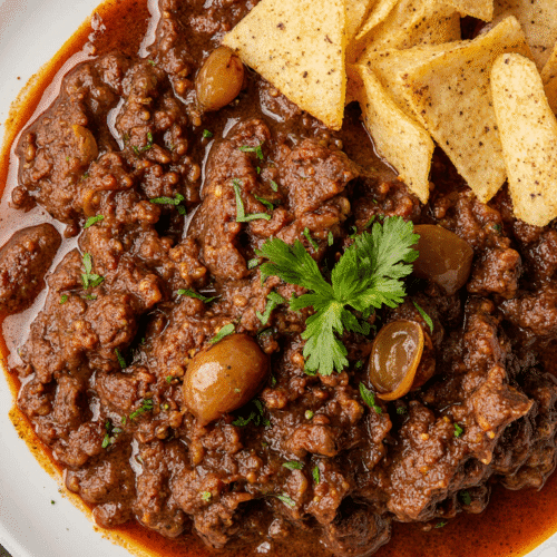 Prato de carne cozida ao molho escuro com azeite, azeitonas e cheiro-verde, servido com nachos crocantes em prato branco. Receita brasileira saborosa e encorpada.